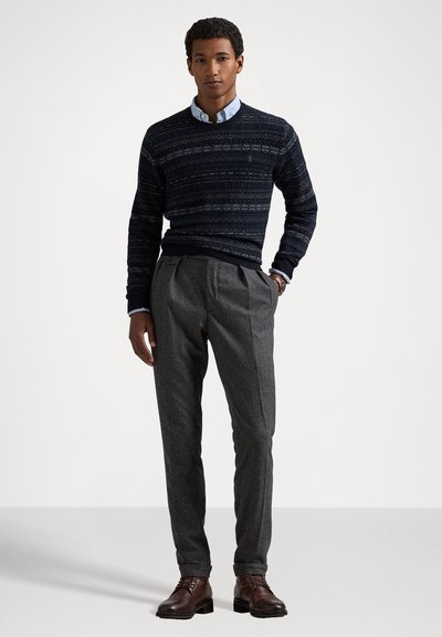 Man wearing a patterned navy sweater over a light blue shirt, gray pleated trousers, and brown leather boots, standing against a plain background.