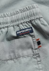 Ceinture en tissu gris avec élastique, arborant une étiquette bleue portant l'inscription "SUPERDRY" et une petite étiquette latérale avec des bandes rouges, blanches et noires.