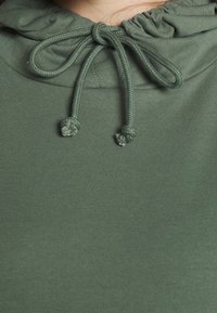 Sudadera con capucha de algodón verde, con cuello con cordón ajustable, tejido fruncido en el cuello y textura visible en la superficie.