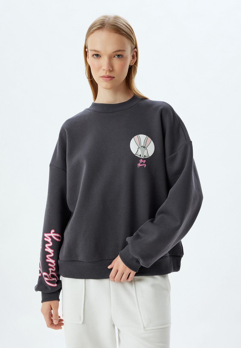 Sudadera gris oscura con gráfico redondo de Bugs Bunny en el pecho; texto en cursiva rosa en la manga; tela suave, corte relajado, cuello y puños acanalados.