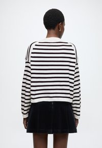 Svītrains džemperis krēmkrāsā un tumši brūnā krāsā ar īsu dizainu, kombinēts ar melnu kordurojas svārku, kuram ir krokas un rāvējslēdzēja detaļa.