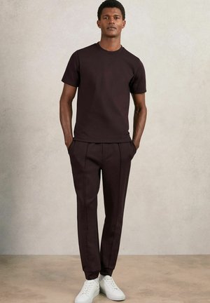 Tricou scurt cu mânecă, de culoare maro închis, asortat cu pantaloni ajustați de culoare maro închis și adidași albi, având un design simplu și o croială lejeră.