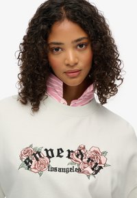 Felpa grigia chiara con rose ricamate e testo. Sotto, una camicia collare rosa e bianca a righe è visibile al collo.