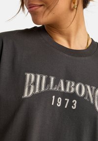 Camiseta de algodón gris oscuro con un gran gráfico de estilo vintage "BILLABONG 1973" en impresión blanca desgastada. Cuello redondo y mangas cortas.