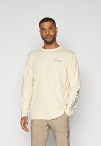 Camicia a maniche lunghe di un leggero color crema, realizzata in tessuto morbido, con un piccolo logo sul petto e un logo più grande lungo la manica sinistra.