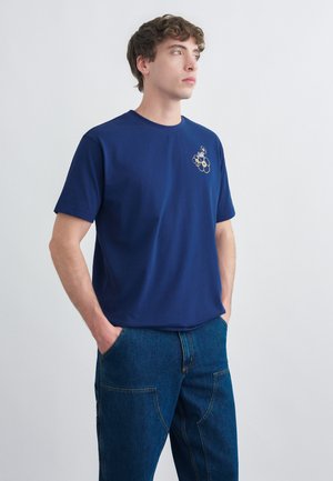 Jeune homme portant un t-shirt bleu marine avec un petit motif d'engrenage beige sur la poitrine et un pantalon en denim bleu, les mains dans les poches, regardant vers la droite.