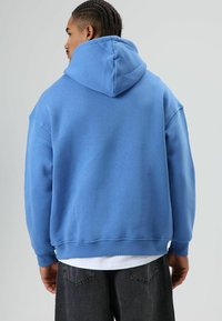Blauwe hooded trui met een ontspannen pasvorm, zachte textuur, geribbelde manchetten en zoom, gedragen over een wit shirt met zwarte spijkerbroek.