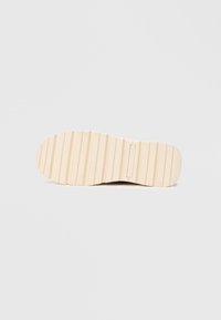 Semelle de chaussure avec une texture nervurée, en caoutchouc. Couleur beige clair avec un détail de logo subtil, forme plate et arrondie.