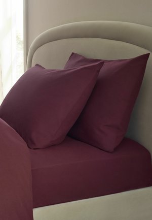 Zwei dunkelviolette Kissen lehnen an einem hellbeigen gebogenen Kopfteil an einem Bett mit passender dunkelvioletter Bettwäsche.