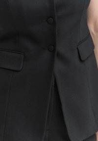 Svart skräddarsydd väst i ett slätt tyg, med två framfickor och tre runda knappar längs knappslån. Rena linjer och en figurnära design.