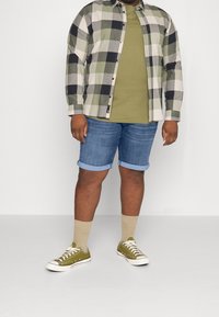 Flannel shirt in green and gray plaid, olive t-shirt, denim shorts, tan socks, and olive sneakers with white soles. Casual outfit.