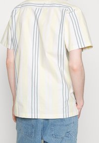 Camicia a maniche corte di un giallo pastello con strisce verticali blu e bianche. Vestibilità leggermente libera e tessuto di cotone morbido. Vista posteriore, stile casual.