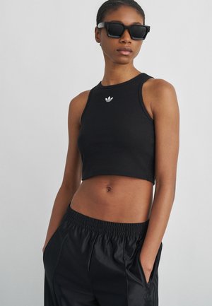 Kobieta w czarnym bezrękawniku typu crop top z logo Adidas, czarne spodnie sportowe i duże czarne okulary przeciwsłoneczne, stoi ze rękami w kieszeniach.