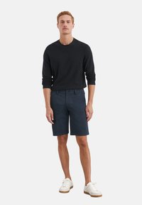 Zwarte gebreide trui en marineblauwe shorts. De shorts hebben een knoopsluiting en voorkeurszakken. De schoenen zijn wit met een lichtbruine zool.