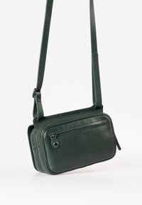 Borsa a tracolla verde in pelle con due scomparti con zip, una tracolla sottile e regolabile, e una texture liscia. Cucitura dettagliata sui bordi.