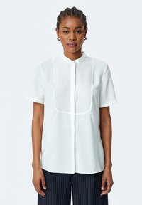 Witte blouse met korte mouwen en hoge kraag, gemaakt van een gladde, lichte stof en met een knoopsluiting aan de voorkant. Subtiele semi-doorzichtige paneeldetails.