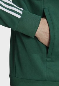 Grön sweatshirt med texturerad tyg, med en sidoficka och vita ränder på ärmen. Muddarna är ribbade för en åtsittande look.