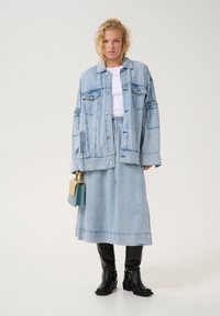 Heleheline teksajakk, mis on üle mõõtme, rinnataskutega, kokku sobitatuna helesinise teksaseelikuga. Mudel hoiab väikest kaherajalist käekotti. Mustad põlvekotid täiustavad välimust.