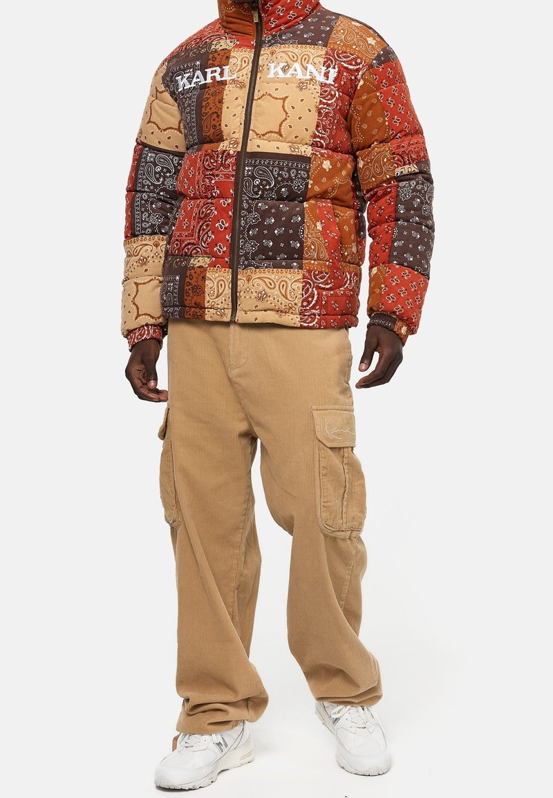 Puffermantel im mehrfarbigen Bandana-Druck mit Schattierungen von Rot, Braun und Beige. Begleitet von beigen Cargo-Hosen und weißen Sneakers.