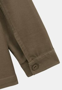 Poignet de chemise à manches longues marron avec fermeture à bouton et couture visible sur un fond gris clair.