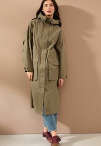 Olivgrüner langer Mantel mit Kapuze, zwei großen Fronttaschen und einer durchgehenden Knopfleiste. Kombiniert mit hellen Jeans und burgunderfarbenen Turnschuhen.