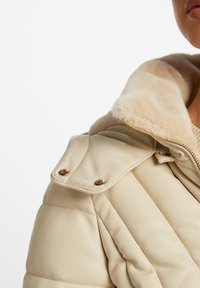 Giacca in pelle beige con colletto soffice, chiusura con zip e dettagli in metallo dorato. Presenta una texture trapuntata e spalle imbottite.