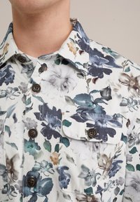Chemise à boutons florale en tissu léger, avec une base blanche ornée de fleurs multicolores en bleu, vert et marron. Comprend une poche sur la poitrine.