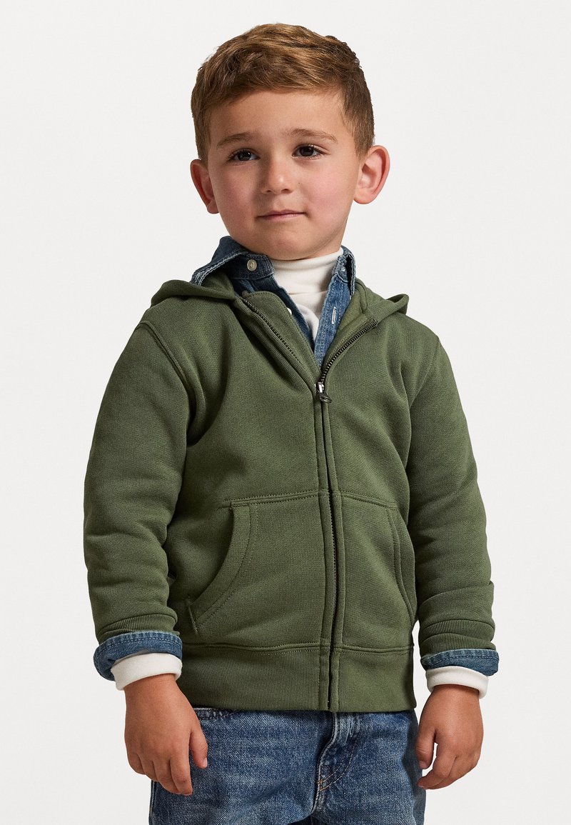 Zelená zipová mikina z měkké tkaniny s předními kapsami, vrstvená přes modrou džínovou košili. Obsahuje žebrované manžety a malý límec.