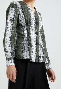 Chemise à col boutonné verte et blanche avec un motif texturé d'arbre de bouleau, comportant quatre boutons noirs et une coupe décontractée. Manches longues avec des poignets.