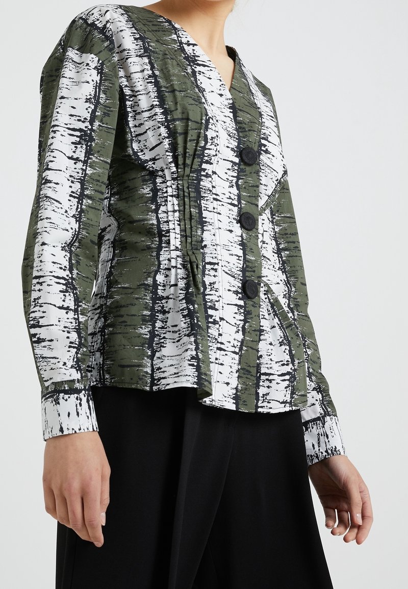 Chemise à col boutonné verte et blanche avec un motif texturé d'arbre de bouleau, comportant quatre boutons noirs et une coupe décontractée. Manches longues avec des poignets.