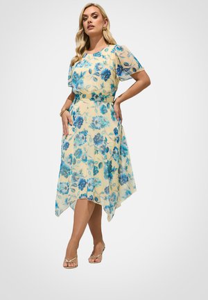 Kvinde iført en gul kjole med blåt blomstermønster, gennemsigtige korte ærmer og asymmetrisk kant, poserer med hænderne på hofterne og gyldne sandaler.