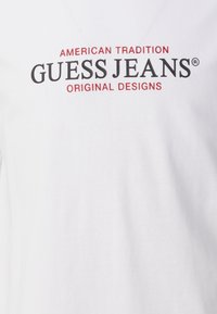 Balts T-kreklis ar melnu un sarkanu drukātu tekstu. Priekšpusē izceltas frāzes "AMERIKĀŅU TRADĪCIJA" un "GUESS JEANS". Izgatavots no kokvilnas auduma.