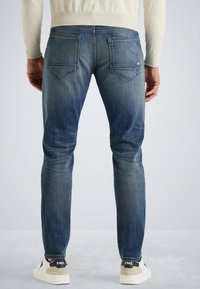 Vaqueros slim fit en denim azul oscuro con un lavado descolorido. Presenta bolsillos traseros y detalles de costura sutiles. Llevados con zapatillas blancas.