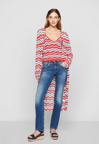 Cárdigan de punto con patrón de zigzag en rojo, azul y blanco sobre una camiseta básica, combinado con jeans de mezclilla azul y sandalias de esparto negras de plataforma.