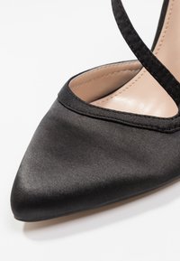 Svart satinhög klack med spetsig tå och delikata korsade remmar. Slät yta med matt finish och en smal klack.