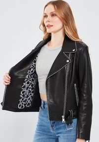 Zwarte leren motorjack met zilvergekleurde studs, ritssluiting zakken en een gevoerde binnenkant van gemusterde stof. Beschikt over een klassiek asymmetrisch model.