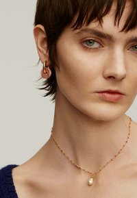 Pendientes de aro dorados con un detalle de disco rosa claro. Un collar de cadena delicado presenta un pequeño colgante de perla. Textura suave y diseño discreto.