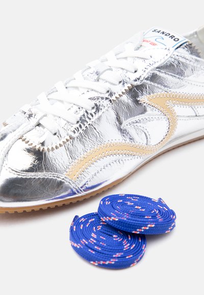 Pantof sport argintiu cu accente bej și șireturi albe, lângă două șireturi albastre încolăcite cu pete roșii și albe pe un fundal alb.