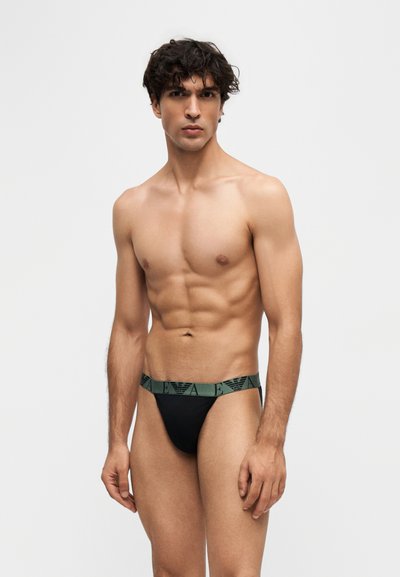 Homme musclé aux cheveux bouclés et foncés portant un slip noir avec une taille verte comportant un logo répété, debout devant un fond clair uni.