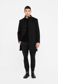 Cappotto di lana nero con collo alto, bottoni frontali e tasche laterali, abbinato a un maglione nero lavorato a maglia e pantaloni neri aderenti, completato da stivali neri.