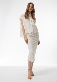 Top de encaje blanco con mangas cortas y falda de encaje a juego, ambas con patrones florales y bordes con forma de concha, combinadas con sandalias nude.