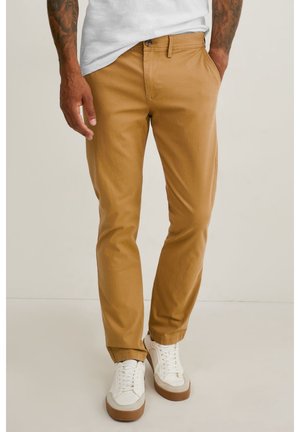 Mann trägt schlanke, enge Chinos in Kamelhaarfarbe, weiße Sneaker mit Gummisohlen und ein weißes T-Shirt, seine tätowierten Arme sind sichtbar, steht vor einem schlichten Hintergrund.