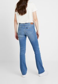 Jeans acampanados azules con una cintura de altura media, elaborados con denim. Presenta una etiqueta de marca en la parte posterior, dos bolsillos traseros y un acabado limpio.