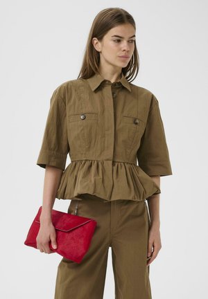 Nő barna, patentos blúzban, peplummal, és hozzá illő nadrágban, kezében élénkvörös borítéktáskával, oldalra néz.