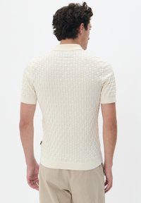 Tejfehér, rövid ujjú pólóing, amely texturált, rácsos mintázattal rendelkezik. Bordás gallérral és szegéllyel rendelkezik. Egyszerű, testhezálló kialakítás.