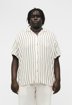 Vyras trumpais dreadlockais dėvi baltą trumpomis rankovėmis marškinius su vertikaliomis rudomis juostomis ir šviesias kelnes, stovintis priešais paprastą foną.