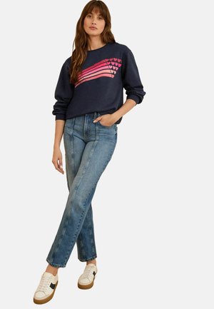 Marineblauer Sweatshirt mit pinkem Herzstreifen-Design, kombiniert mit hochtaillierten blauen Jeans. Das Model trägt weiße Sneakers mit schwarzen Akzenten.