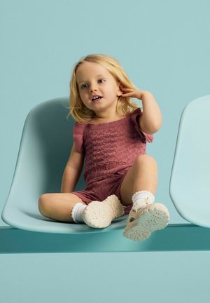 Jeune enfant aux cheveux blonds portant une tenue en tricot rose et des baskets blanches, assis jambes croisées sur une chaise bleu clair sur un fond assorti.