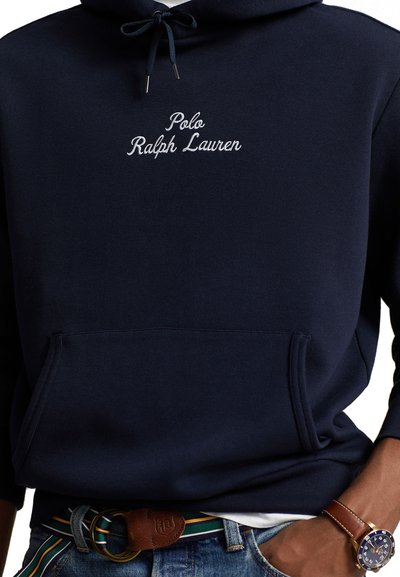 Tamsiai mėlynas medvilnės džemperis su priekinėmis kišenėmis; ant jo baltais siuvinėjimais užrašyti "Polo Ralph Lauren". Tai yra aprangos dalis su džinsais ir ruduo diržu.