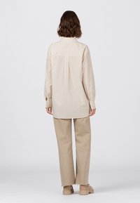 Beige Langarmhemd mit gerader Passform, Rückenfalten und geknöpften Manschetten, kombiniert mit hellbeigen weit geschnittenen Hosen und klobigen beigen Schuhen.